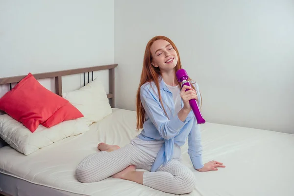 Kızıl saçlı kadın, yatakta mikrofon gibi, pembe yastıklı seks oyuncağı tutuyor.