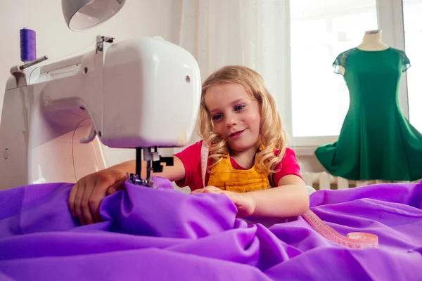 Smiling little blonde girl at the table with sewing machine with manikin — Stock Photo, Image