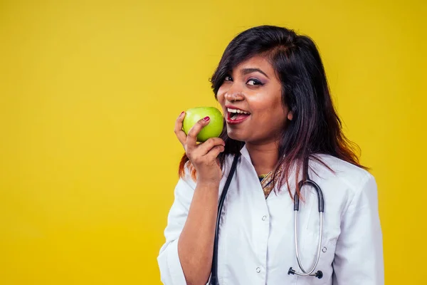Kobieta indyjski młody i piękny blond kobieta ginekolog lekarz za pomocą stetoskopu trzymając zielone jabłko w dłoni w białym płaszczu medycznym na żółtym tle w studio — Zdjęcie stockowe