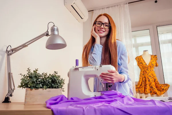 Donna d'affari dai capelli rossi con macchina da cucire e manichino sullo sfondo dello studio. sarto crea una collezione abiti cuce vestiti in officina scrittura idea su appunti di vestiti — Foto Stock