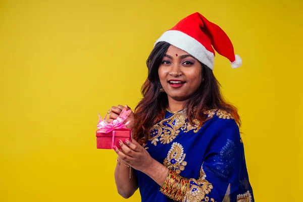 Kvinna porträtt bär traditionella india sari och jul Santa hatt grepp jul gåva gul bakgrund studio — Stockfoto