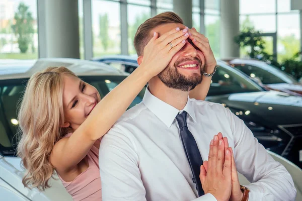 Casal feliz stand na frente do carro novo, homem coberto esposa olhos em showroom.surprised presente automóvel — Fotografia de Stock
