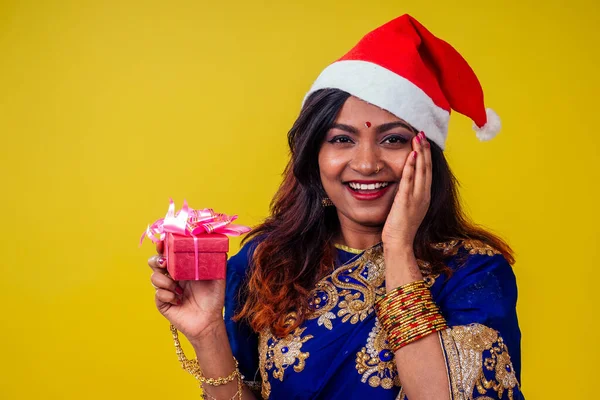 Donna ritratto indossando tradizionale india sari e Natale Santa cappello tenere Natale gifton giallo sfondo studio — Foto Stock