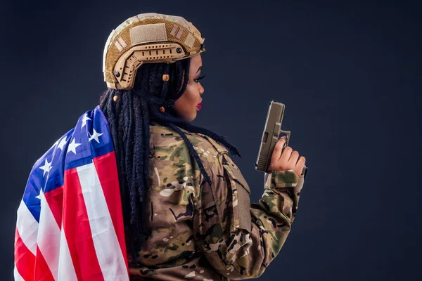 Afro amerykański wojsko latynoski żołnierz w kamuflażu ubrania dredy włosy wieczorem makijaż i duże usta trzymając broń na czarnym tle w studio z amerykańską flagą, Dzień Niepodległości usa 4 lipca — Zdjęcie stockowe