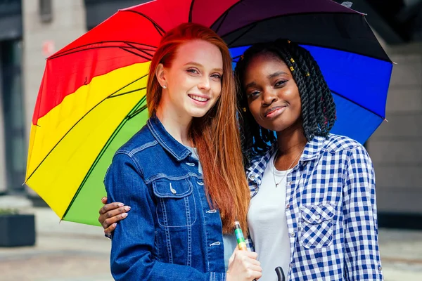 Latino hispânico e ruiva ruiva mulheres segurando guarda-chuva arco-íris na rua — Fotografia de Stock