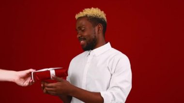 Kırmızı stüdyo arka planında beyaz tişörtlü Latin bir adam ve küçük hediye kutusu var.