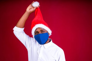 Kırmızı stüdyo arka planında maske takan, Noel Baba şapkalı Afro-Amerikan bir adam.
