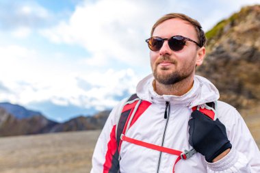 Genç sakallı, beyaz kabarık ceketli bir adam bir dağın yanında duruyor.
