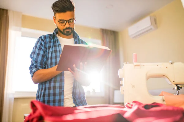 indian tailor asian man designer sewing clothes on sewing machine next to laptop home apartment workplace