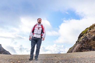Genç sakallı, beyaz kabarık ceketli bir adam bir dağın yanında duruyor.