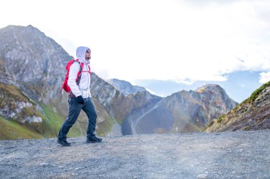Genç sakallı, beyaz kabarık ceketli bir adam bir dağın yanında duruyor.