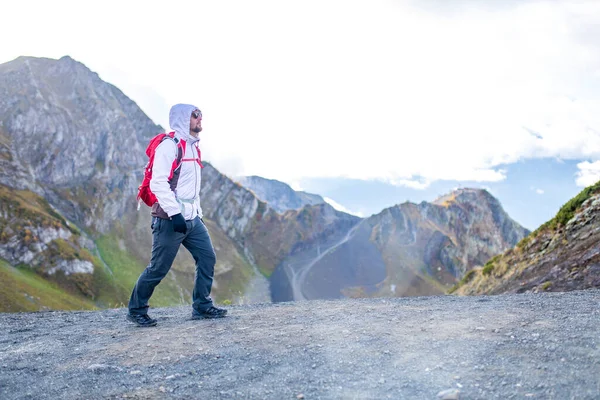 Genç sakallı, beyaz kabarık ceketli bir adam bir dağın yanında duruyor.