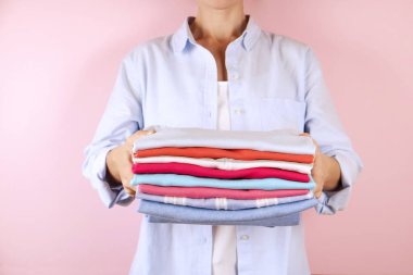 Mükemmel katlanmış çok renkli gömlek yığını tutan mavi pamuklu gömlek giyen genç kadın yakın çekim. Erkek w / ellerinin soluk pembe bir arka plan üzerinde farklı renk giyim yığını. Çamaşırhane kavramı.