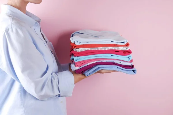 近身身穿蓝棉布衬衫的年轻女子抱着一叠完全折叠的多色衬衣 堆不同颜色的衣服在她的手在淡粉红色的背景 洗衣概念 — 图库照片