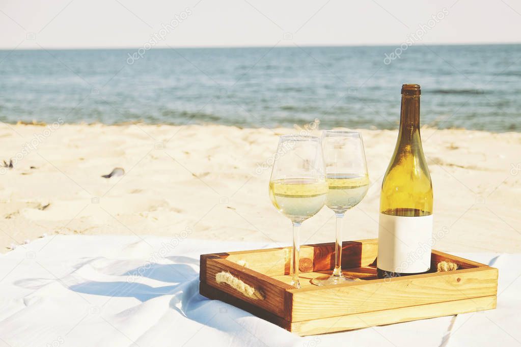 Romantic dinner composition with expensive bottle of white wine, blank label and two glasses on wooden tray at the white sandy beach, ocean. Sea view. Close up, background, copy space.