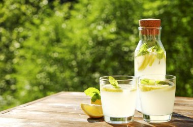 Limonata sürahi ve iki bardak soğuk ferahlatıcı narenciye içecek limon ve nane ile yaprakları kahverengi grunged ahşap masa, ülke tarafı yeşillik, yeşil arka plan üzerinde buz dolu. Yakın çekim, alanı Kopyala.