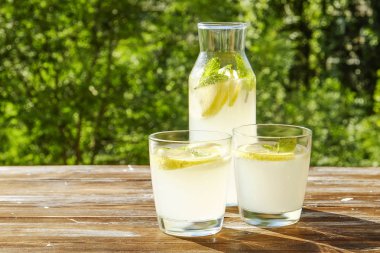 Limonata sürahi ve iki bardak soğuk ferahlatıcı narenciye içecek limon ve nane ile yaprakları kahverengi grunged ahşap masa, ülke tarafı yeşillik, yeşil arka plan üzerinde buz dolu. Yakın çekim, alanı Kopyala.