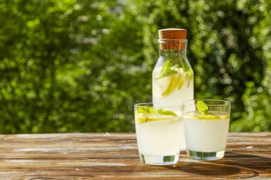 Limonata sürahi ve iki bardak soğuk ferahlatıcı narenciye içecek limon ve nane ile yaprakları kahverengi grunged ahşap masa, ülke tarafı yeşillik, yeşil arka plan üzerinde buz dolu. Yakın çekim, alanı Kopyala.