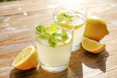 İki bardak nane ile buz soğuk ferahlatıcı limonata içecek tam güneş ışığı altında kahverengi grunged ahşap masa üzerinde olgun organik limon, bütün ve yarıya dilim bırakır. Arka plan, kopya alanı, yakın çekim.