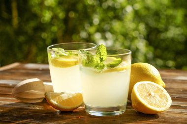 İki bardak buz dolu ferahlatıcı soğuk limonata içecek nane w / güneş ışığında yaprakları kahverengi grunged ahşap masa, meyva suyu sıkacağı, olgun organik limon, ülke tarafı yeşillik arka plan üzerinde. Uzay kopyalayın, yakın çekim.