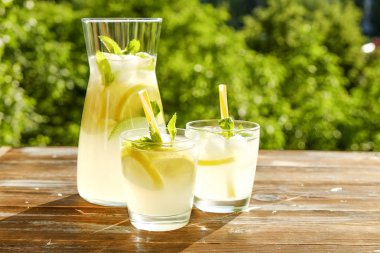 Limonata sürahi ve iki bardak soğuk buz, limon dilimleri, nane yaprakları ve kahverengi grunged ahşap masa, ülke tarafı yeşillik arka plan üzerinde payet ile narenciye içecek yenileniyor. Yakın çekim, alanı Kopyala.