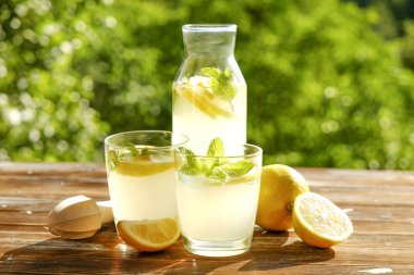 Limonata sürahi, meyva suyu sıkacağı, iki bardak limon dilimleri ve nane yaprakları kahverengi grunged ahşap masa, ülke tarafı yeşillik arka plan ile buz soğuk ferahlatıcı narenciye içecek dolu. Yakın çekim, alanı Kopyala.
