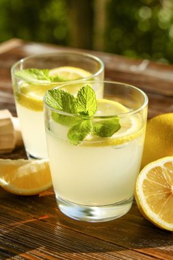 İki bardak buz dolu ferahlatıcı soğuk limonata içecek nane w / güneş ışığında yaprakları kahverengi grunged ahşap masa, meyva suyu sıkacağı, olgun organik limon, ülke tarafı yeşillik arka plan üzerinde. Uzay kopyalayın, yakın çekim.