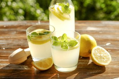 Limonata sürahi, meyva suyu sıkacağı, iki bardak limon dilimleri ve nane yaprakları kahverengi grunged ahşap masa, ülke tarafı yeşillik arka plan ile buz soğuk ferahlatıcı narenciye içecek dolu. Yakın çekim, alanı Kopyala.