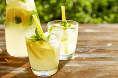 Limonata sürahi ve iki bardak soğuk buz, limon dilimleri, nane yaprakları ve kahverengi grunged ahşap masa, ülke tarafı yeşillik arka plan üzerinde payet ile narenciye içecek yenileniyor. Yakın çekim, alanı Kopyala.