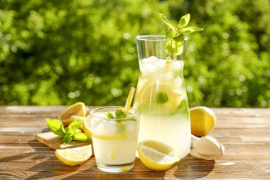 Buzlu limonata sürahi, meyve sıkacağı ve limon dilimleri, nane yaprakları ve kahverengi grunged ahşap masa, ülke tarafı yeşillik arka plan üzerinde sarı payet ile soğuk narenciye içecek bir bardak. Yakın çekim, alanı Kopyala.