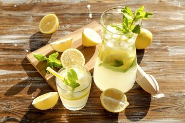 Buzlu limonata sürahi, ahşap meyve sıkacağı ve limon dilimleri, nane yaprakları ve kahverengi grunged Ahşap dokulu masa güneşli, arka plan sarı saman ile soğuk narenciye içecek bir bardak. Yakın çekim, alanı Kopyala.