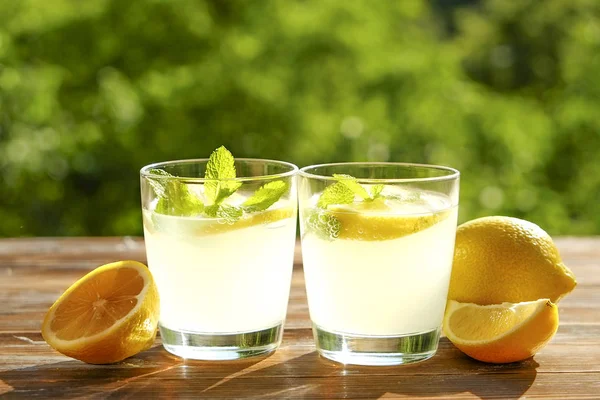 İki bardak buz soğuk ferahlatıcı limonata içecek nane w / dolu kahverengi grunged ahşap masa üzerinde olgun organik limon, ülke tarafı yeşillik arka plan dilim güneş ışığı altında bırakır. Uzay kopyalayın, yakın çekim.