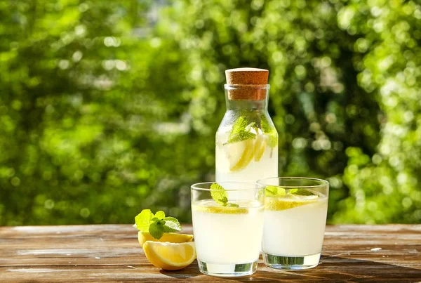 Limonata sürahi ve iki bardak soğuk ferahlatıcı narenciye içecek limon ve nane ile yaprakları kahverengi grunged ahşap masa, ülke tarafı yeşillik, yeşil arka plan üzerinde buz dolu. Yakın çekim, alanı Kopyala.