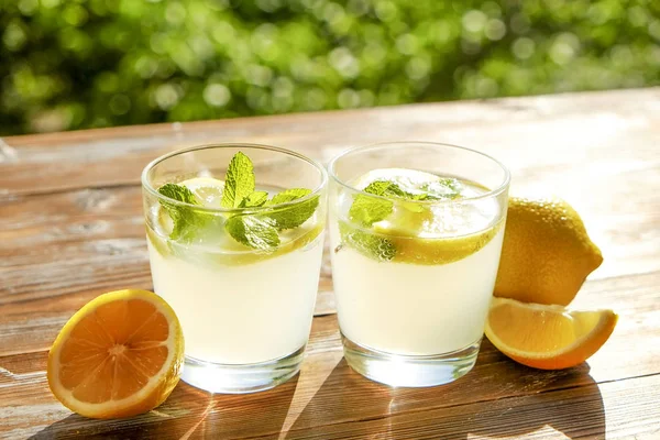 İki bardak buz soğuk ferahlatıcı limonata içecek nane w / dolu kahverengi grunged ahşap masa üzerinde olgun organik limon, ülke tarafı yeşillik arka plan dilim güneş ışığı altında bırakır. Uzay kopyalayın, yakın çekim.