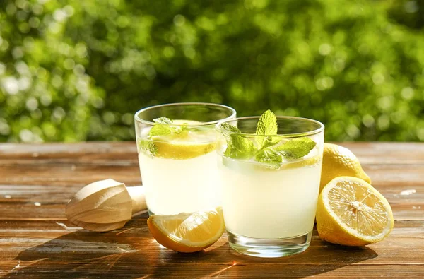 İki bardak buz soğuk ferahlatıcı limonata içecek nane w / dolu kahverengi grunged ahşap masa üzerinde olgun organik limon, ülke tarafı yeşillik arka plan dilim güneş ışığı altında bırakır. Uzay kopyalayın, yakın çekim.