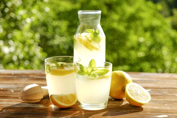 Limonata sürahi, meyva suyu sıkacağı, iki bardak limon dilimleri ve nane yaprakları kahverengi grunged ahşap masa, ülke tarafı yeşillik arka plan ile buz soğuk ferahlatıcı narenciye içecek dolu. Yakın çekim, alanı Kopyala.