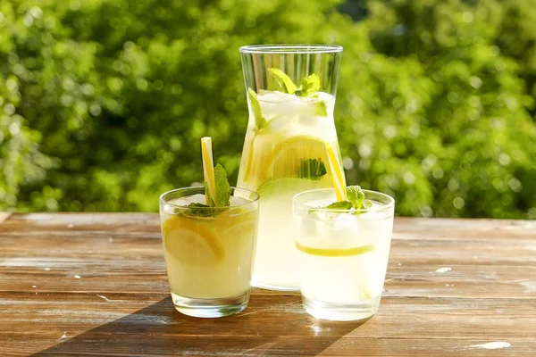 Limonata sürahi ve iki bardak soğuk buz, limon dilimleri, nane yaprakları ve kahverengi grunged ahşap masa, ülke tarafı yeşillik arka plan üzerinde payet ile narenciye içecek yenileniyor. Yakın çekim, alanı Kopyala.