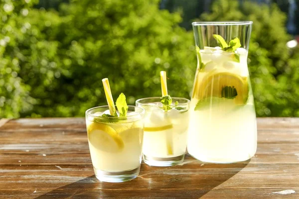 Limonata sürahi ve iki bardak soğuk buz, limon dilimleri, nane yaprakları ve kahverengi grunged ahşap masa, ülke tarafı yeşillik arka plan üzerinde payet ile narenciye içecek yenileniyor. Yakın çekim, alanı Kopyala.