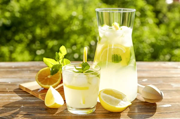 Buzlu limonata sürahi, meyve sıkacağı ve limon dilimleri, nane yaprakları ve kahverengi grunged ahşap masa, ülke tarafı yeşillik arka plan üzerinde sarı payet ile soğuk narenciye içecek bir bardak. Yakın çekim, alanı Kopyala.