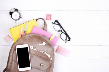 Hipster gri deri sırt çantası, okul malzemeleri, boş ekran cep telefonu, kulaklık, pembe ve sarı defter, Gözlük, mini çalar saat dolu. Geri okul kavramı. Yakın çekim, uzay, düz yatıyordu Kopyala.