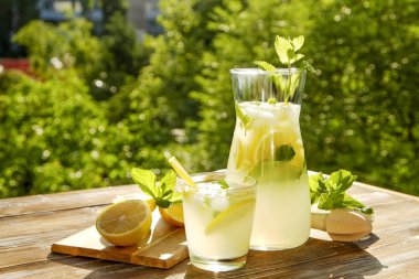 Buzlu limonata sürahi, meyve sıkacağı ve limon dilimleri, nane yaprakları ve kahverengi grunged ahşap masa, ülke tarafı yeşillik arka plan üzerinde sarı payet ile soğuk narenciye içecek bir bardak. Yakın çekim, alanı Kopyala.