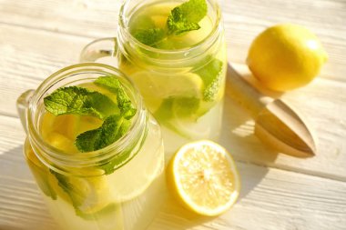 Two mason jar glasses of homemade refreshing lemonade, slices of organic ripe lemon, whole and halved, mint leaves, juicer, muddler, squeezer on a rustic white wooden background. Close up, top view. clipart