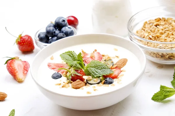 Serving option of granola bowl with mix of nuts, cereals, fruits and berries, greek yogurt. Healthy vegetarian breakfast, organic strawberry, blueberry, mint, almond. Close up, top view, background.