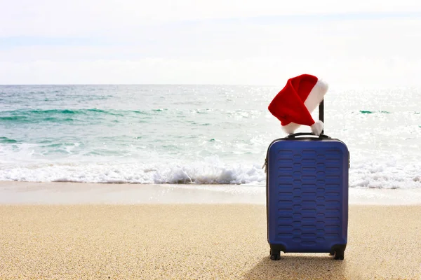 Blue hardshell carry-on roller luggage standing on sandy beach with ocean waves and clear sky on background. Hard plastic suitcase on wheels, santa claus hat on extended handle. Copy space, close up.