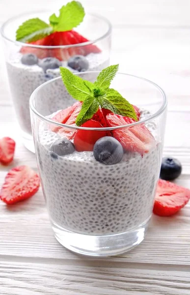 Two portions of chia pudding with vegan almond milk, blueberry & strawberry, mint, served in glasses. Healthy vegetarian breakfast, seeds, berries, greek yogurt, spoon. Background, close up, top view.