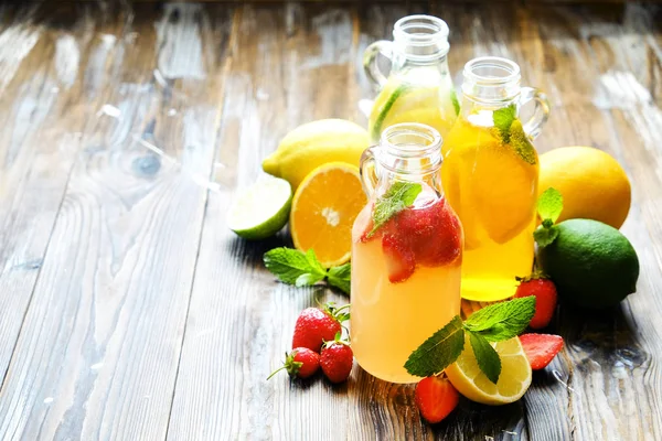 Three vintage bottles of fresh ice cold lemonade, different taste drinks w/ lemon, orange, grapefruit, lime, mint leaves & strawberry on grunged wooden table background. Top view, copy space, close up