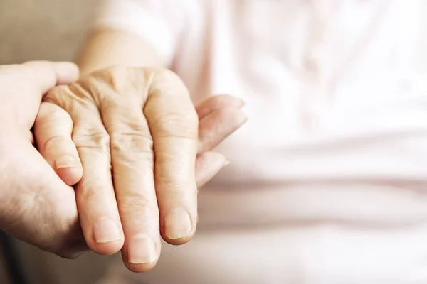 Mature female in elderly care facility gets help from hospital personnel nurse. Senior woman w/ aged wrinkled skin & care giver, hands close up. Grand mother everyday life. Background, copy space.