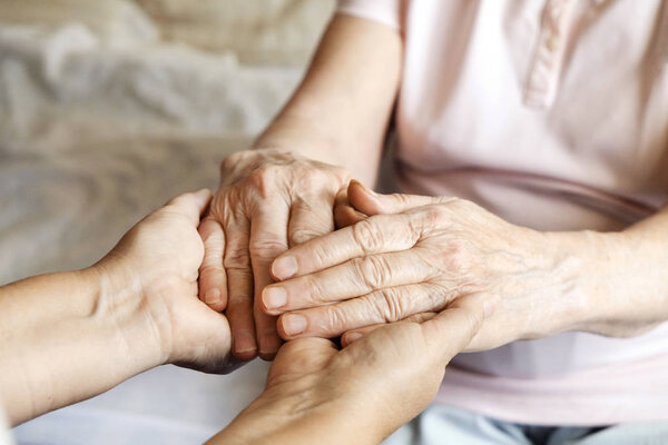 Mature female in elderly care facility gets help from hospital personnel nurse. Senior woman w/ aged wrinkled skin & care giver, hands close up. Grand mother everyday life. Background, copy space.