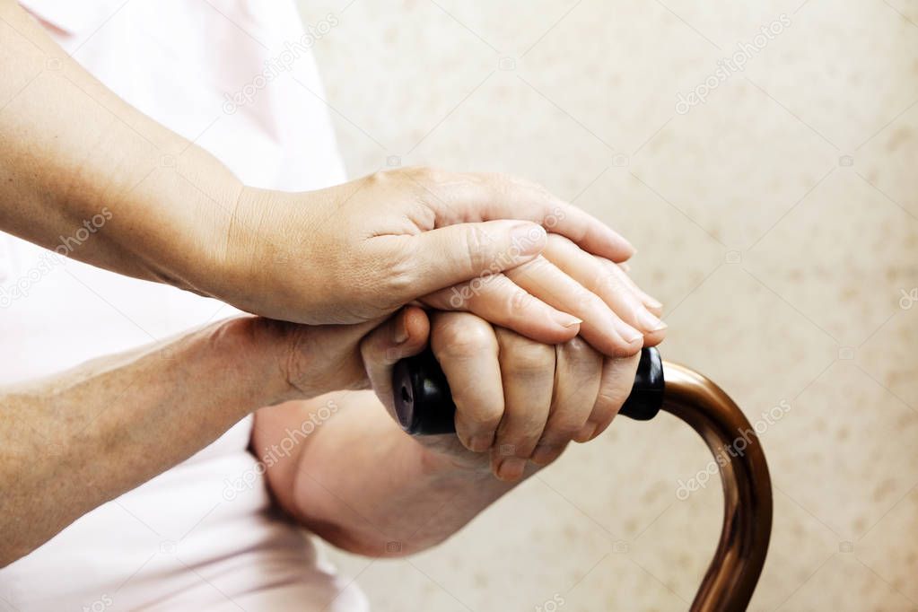 Senior woman holding quad cane handle in elderly care fecility. Hospital nurse comforting mature female in nursing home, sitting with walking stick. Background, close up on hands w/ wrinkled skin.