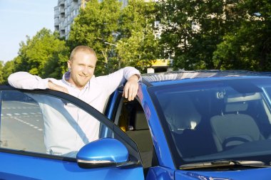 Genç adam onun yeni Karayip mavi araba yaslanmış beyaz gömlek gülümseyen portresi. Otomobil memnun yüz ifadesi, kentsel arka plan ile yakın duran yakışıklı sarışın iş adamı. Kopya alanı